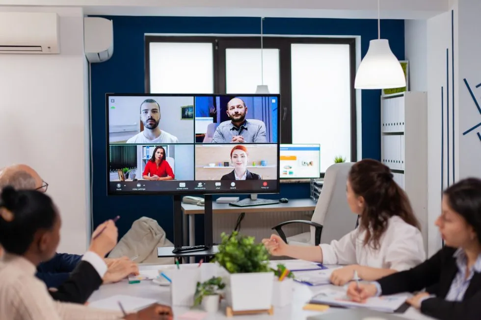People in an office conference room attending a video call with five participants shown on a large screen.